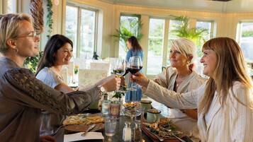 Un grupo de personas brindando con copas de vino en una mesa llena de diversos platos, incluyendo una pizza y una bandeja de mariscos, en una sala con ventanas amplias y plantas.