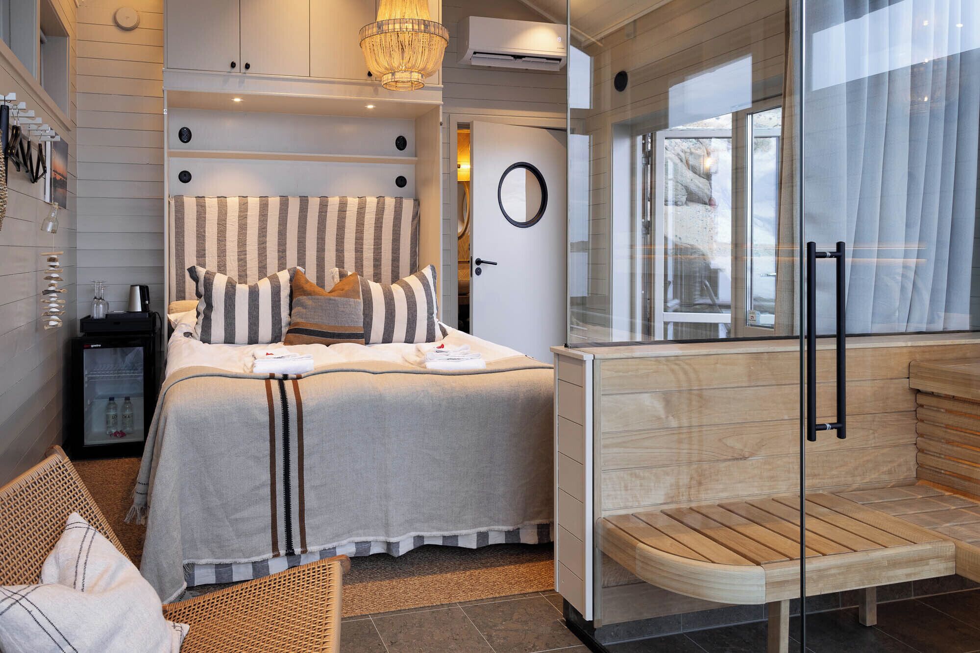 A modern hotel room with a bed, a glass shower enclosure, a wooden bench, and a view of the outdoors.