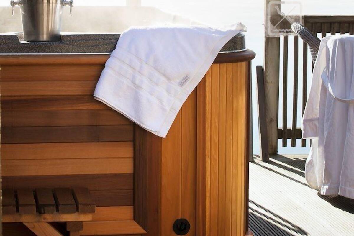 A wooden hot tub with a towel draped over it, a pair of slippers, and a pot on the side.