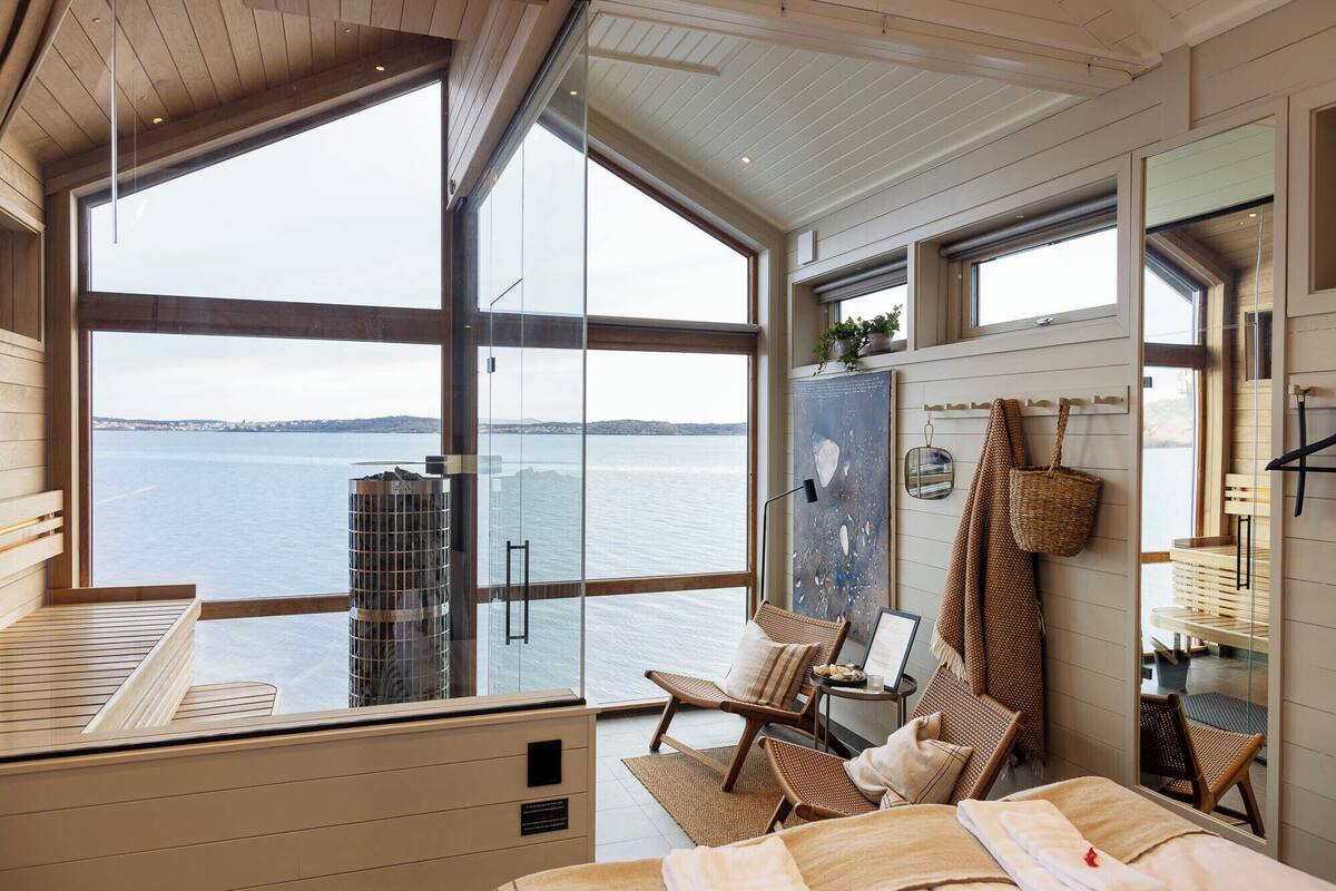 A room with large windows overlooking a body of water, a bed, a chair, a small table, and a wall-mounted rack with items.