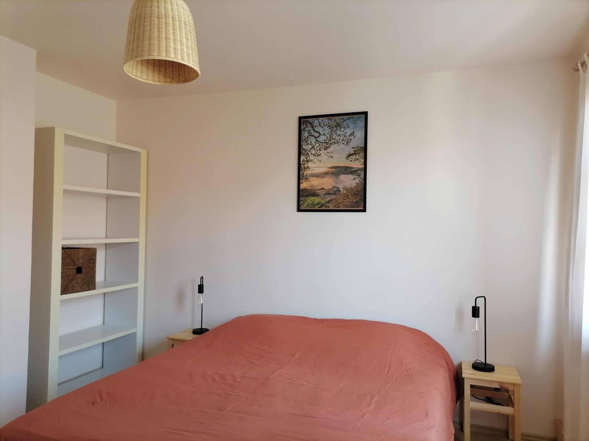Une chambre à coucher avec un lit, une étagère blanche, un tableau représentant un paysage encadré et deux lampes de chevet.