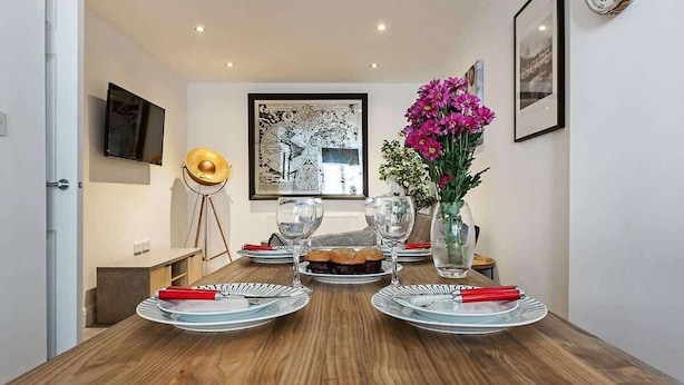 Uma área de refeições com uma mesa de madeira posta para quatro pessoas, uma obra de arte emoldurada na parede, um relógio e um vaso com flores.