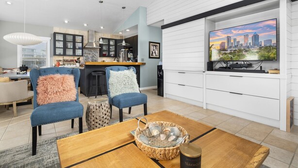 Una sala de estar moderna con televisor mostrando un paisaje urbano, una zona de cocina y un espacio comedor con mesa y sillas de madera.
