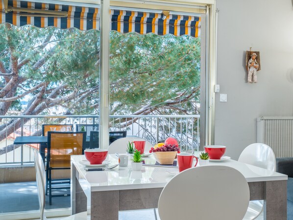 A dining area with a table set for two, chairs, and a view of trees through large windows. - 2-Room Apartment with Terrace, Sea View (Cannes)