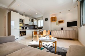 A modern living room with a sofa, a dining area with chairs and a table, a kitchen with cabinets and appliances, a TV, and a clock on the wall. - 2-Bedroom apartment in Grand Hotel (Cannes)