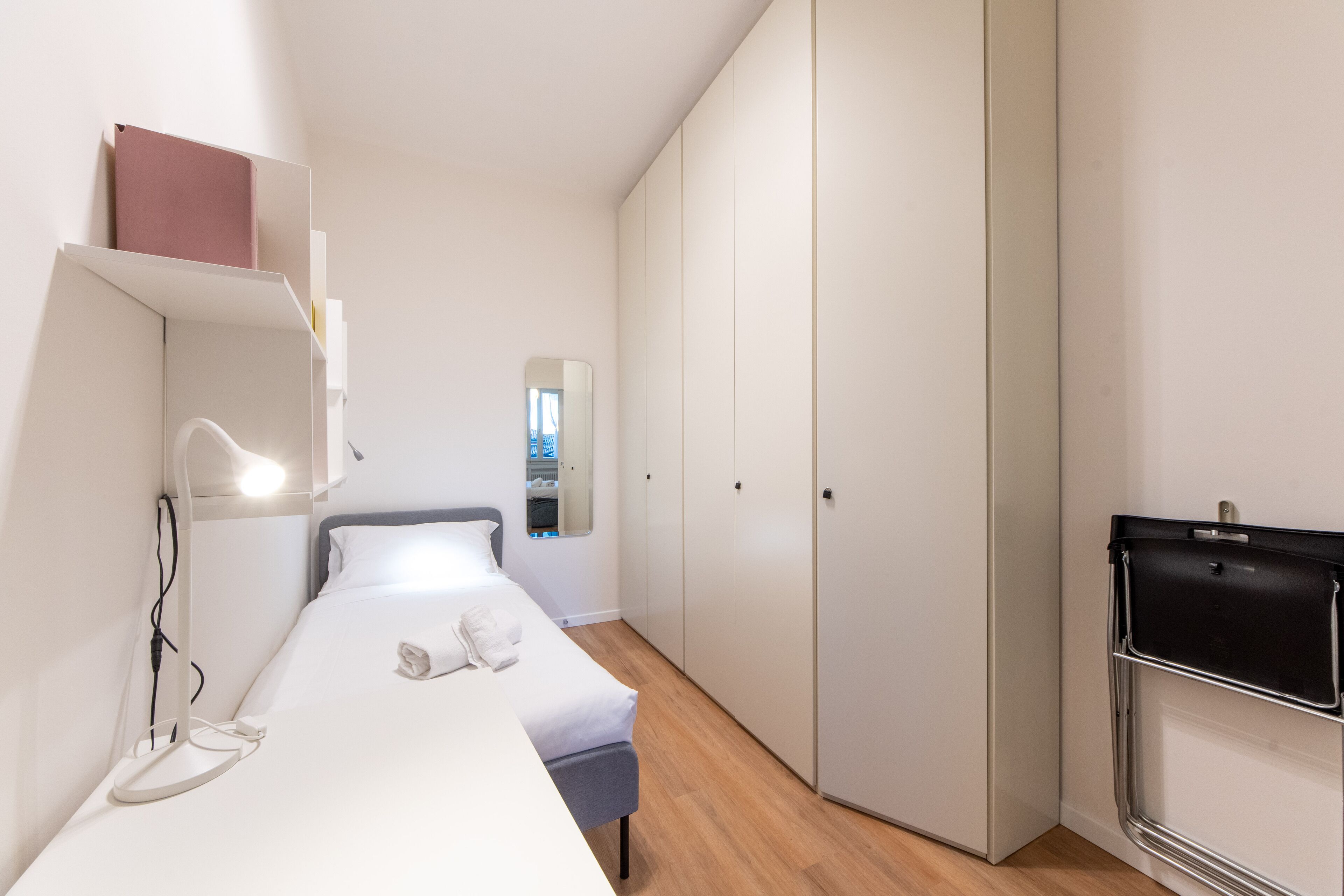 Quarto de hotel compacto com cama, mesa de cabeceira, luminária, guarda-roupa e uma pequena escrivaninha com cadeira.