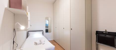 A compact hotel room with a bed, bedside table, lamp, wardrobe, and a small desk with a chair.