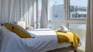 A hotel room with a bed, curtains, and a view of the city through large windows.