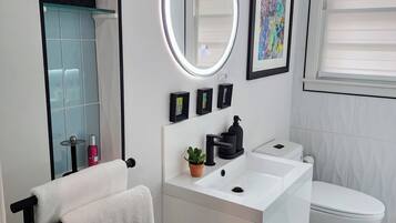 Un baño moderno con un tocador blanco, un espejo redondo con iluminación indirecta, un inodoro y un lavamanos. Hay cuadros enmarcados en la pared y una planta en maceta sobre el tocador.