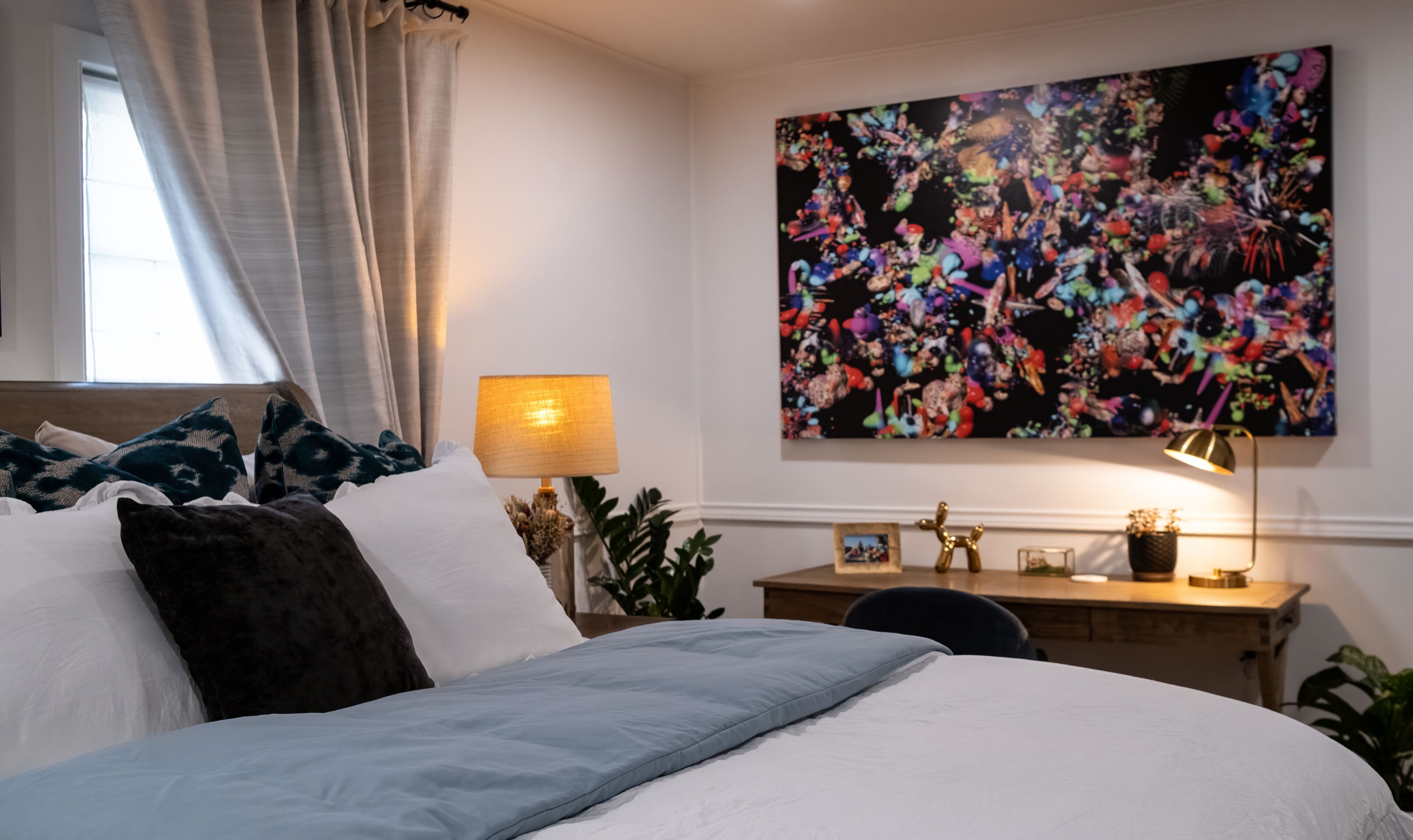A bedroom with a bed, pillows, a desk, a lamp, and a large abstract painting on the wall.
