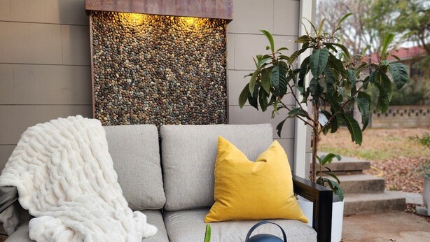 Un espace de détente extérieur moderne, avec un canapé gris, un plaid blanc et un coussin jaune. Sur la table, on trouve deux tasses blanches et une plante en pot.
