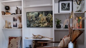 Un salon avec un grand tableau, des étagères ornées d’objets décoratifs, une chaise en bois et une petite table sur laquelle est posé un vase.