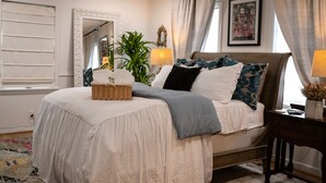 Un lit bien fait, avec une couette blanche, un plaid bleu et plusieurs oreillers. On y trouve un panier en osier, une table de chevet avec une lampe et une plante en pot près de la fenêtre.