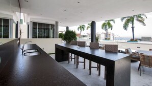 Een modern buitenrestaurant met een lange zwarte tafel, comfortabele zitplaatsen en uitzicht op palmbomen en gebouwen.