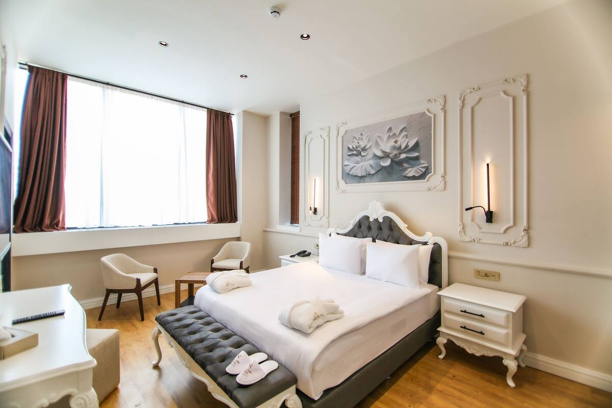 A hotel room with a large bed, a desk, a chair, a bench, and a bedside table.