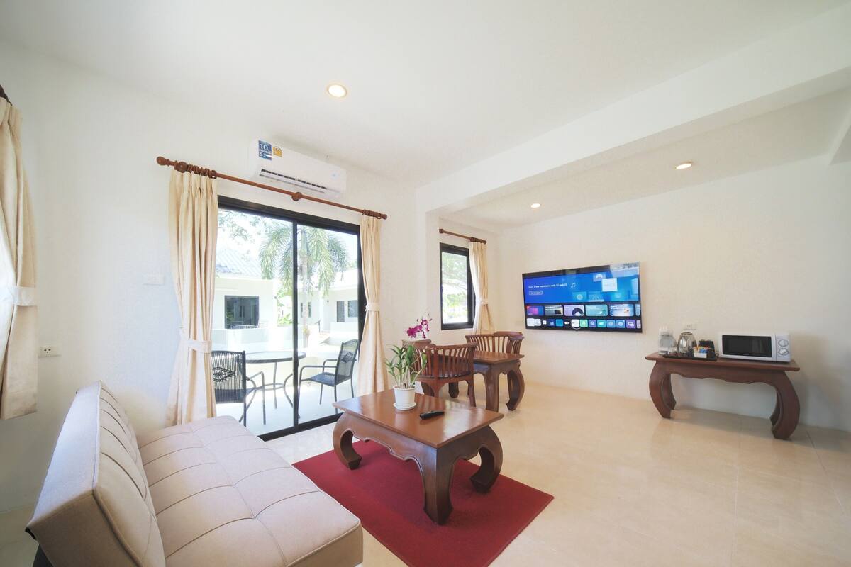 A living room with a sofa, dining table, chairs, a TV mounted on the wall, and an air conditioning unit.