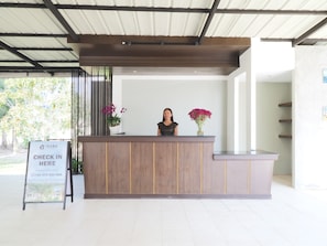 A reception desk with a woman standing behind it, a signboard with contact information, and potted plants. - Isara Nai Yang Resort (Sa Khu)