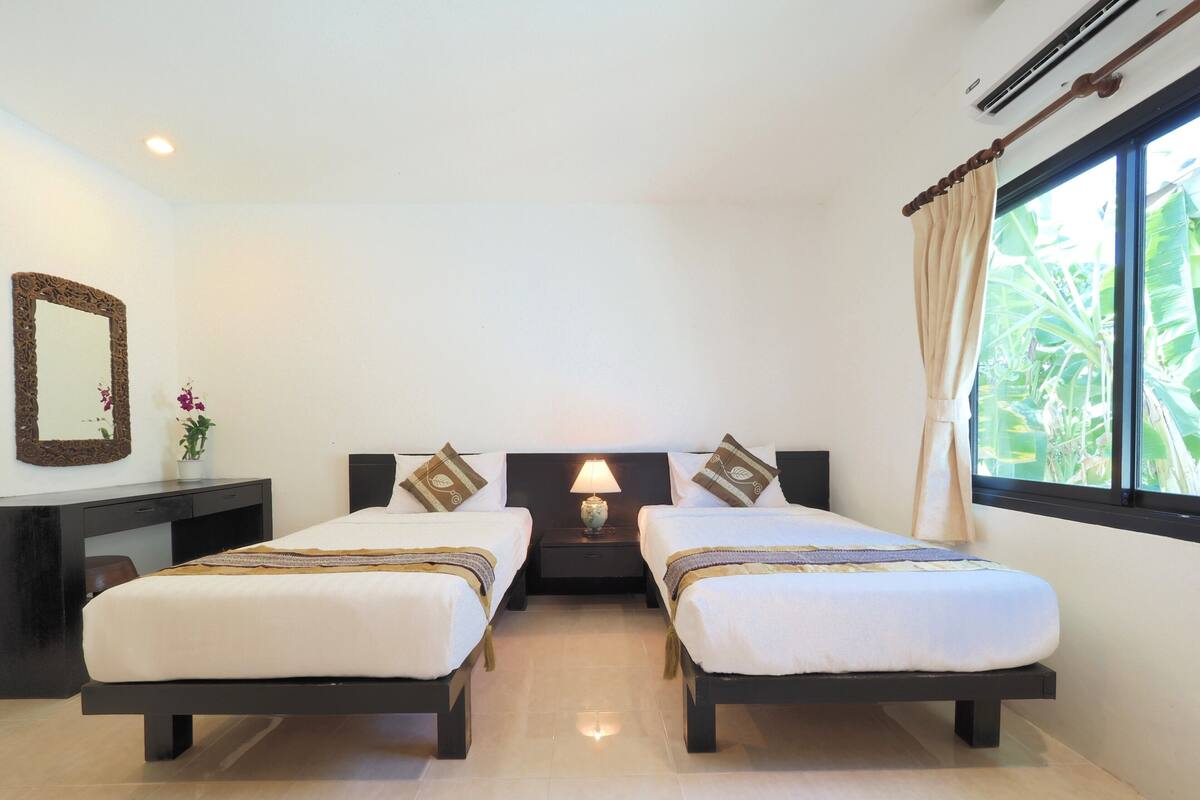 A hotel room with two beds, a nightstand, a desk, a mirror, and a window with a view of greenery.