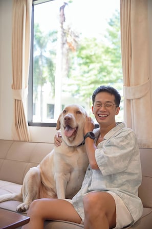 Uma pessoa sentada num sofá com um cão, uma janela com cortinas e vista para o verde lá fora.