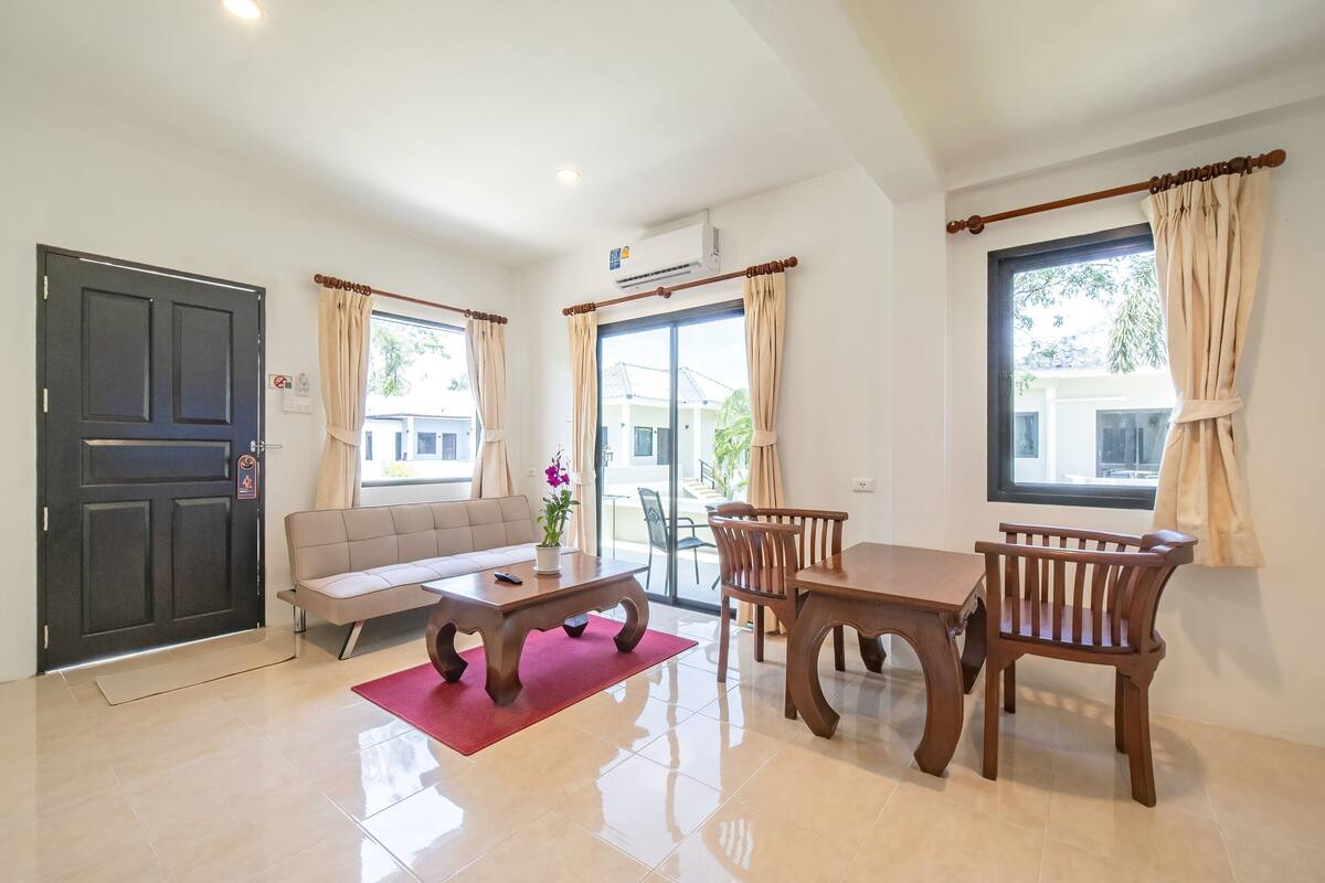 A spacious living area with a sofa, dining table, and chairs, a large window with curtains, and a door with a security lock.