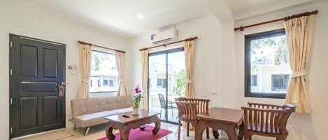 A spacious living area with a sofa, dining table, and chairs, a large window with curtains, and a door with a security lock.