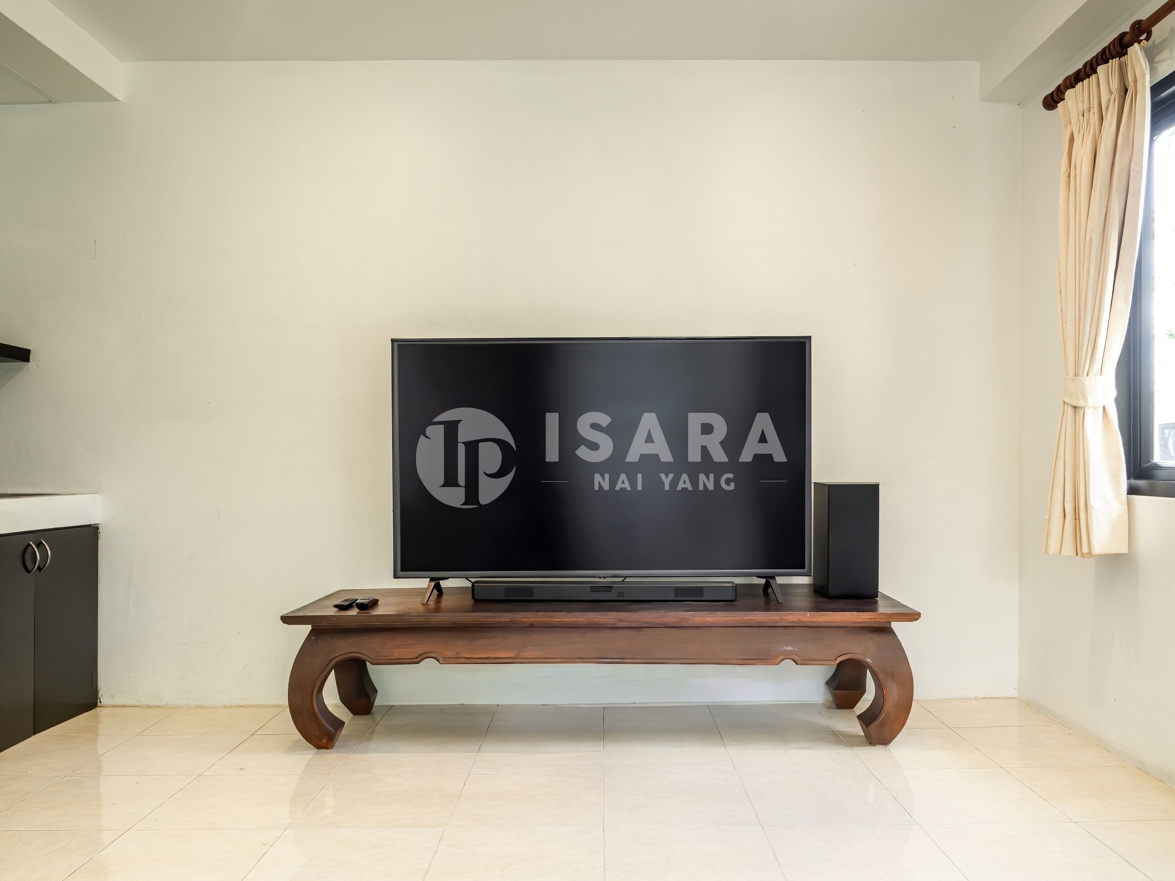 A flat-screen TV on a wooden stand with a logo on the screen, a speaker, and a window with curtains.