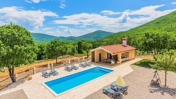 Area piscina con lettini e un edificio dal tetto rosso, immersa nel verde e tra colline.