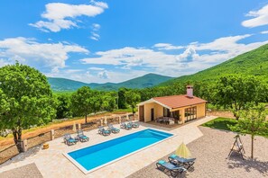 A pool area with lounge chairs and a building with a red roof, surrounded by greenery and hills.