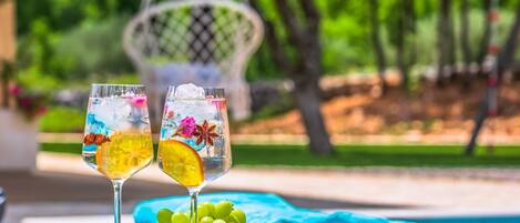 Two glasses of a colorful drink with ice and fruit garnishes, a bowl of green grapes, and a lime half on a table by a poolside with a chair and greenery in the background.