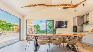 Un'ampia sala da pranzo con tavolo in legno e sedie in vimini, una grande finestra vista su vegetazione e un edificio, e una cucina moderna con televisore a schermo piatto.