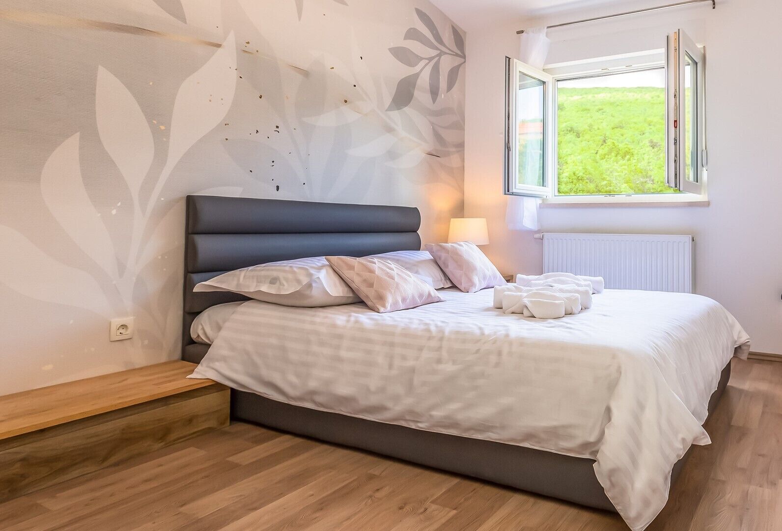 A neatly made bed with white linens and multiple pillows, a dark headboard, and a window with a view of greenery.