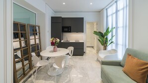 Cocina moderna con armarios oscuros, encimera blanca y microondas integrado. Hay una mesa de comedor con sillas blancas y un jarrón con flores. Una planta verde grande está en la esquina, y un sofá verde con un cojín marrón está apoyado en la pared.