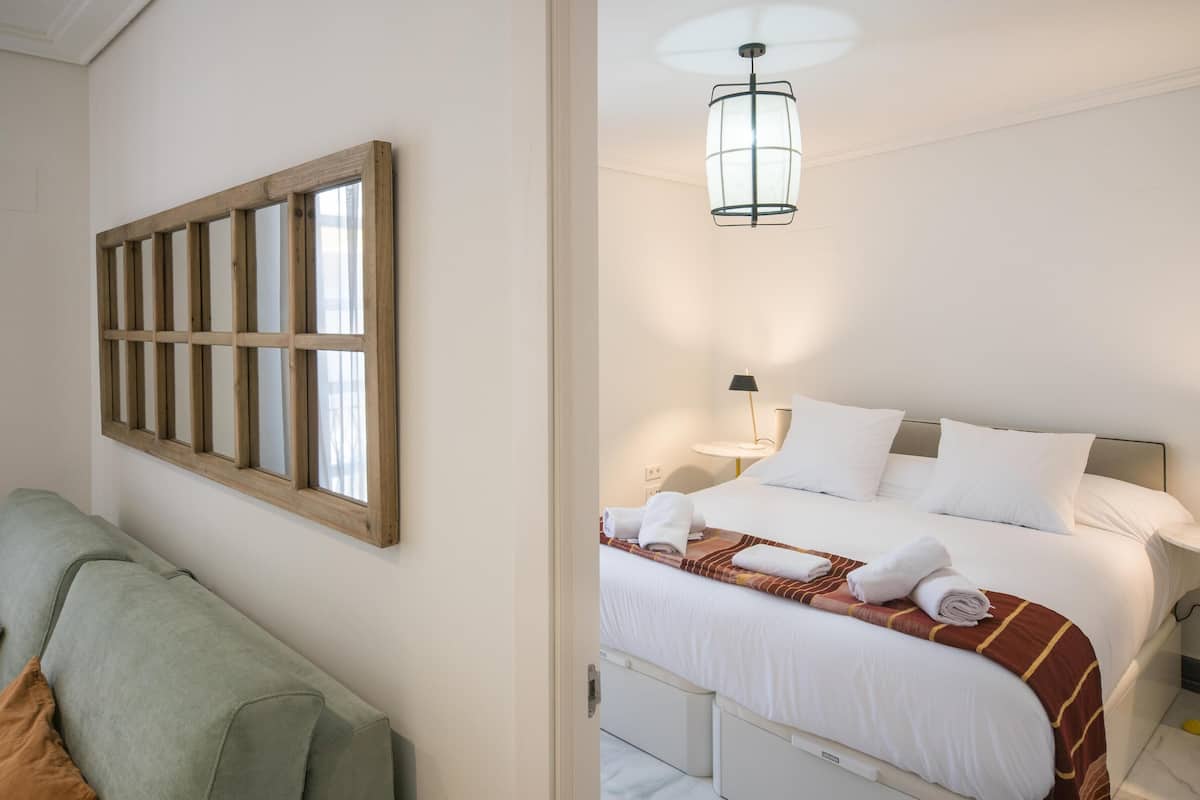 A hotel room with a bed, a nightstand, a lamp, a window with wooden blinds, and a green sofa.