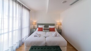 Una cama doble con ropa de cama blanca, dos mesitas de noche con lámparas, un banco verde y cortinas de gasa.