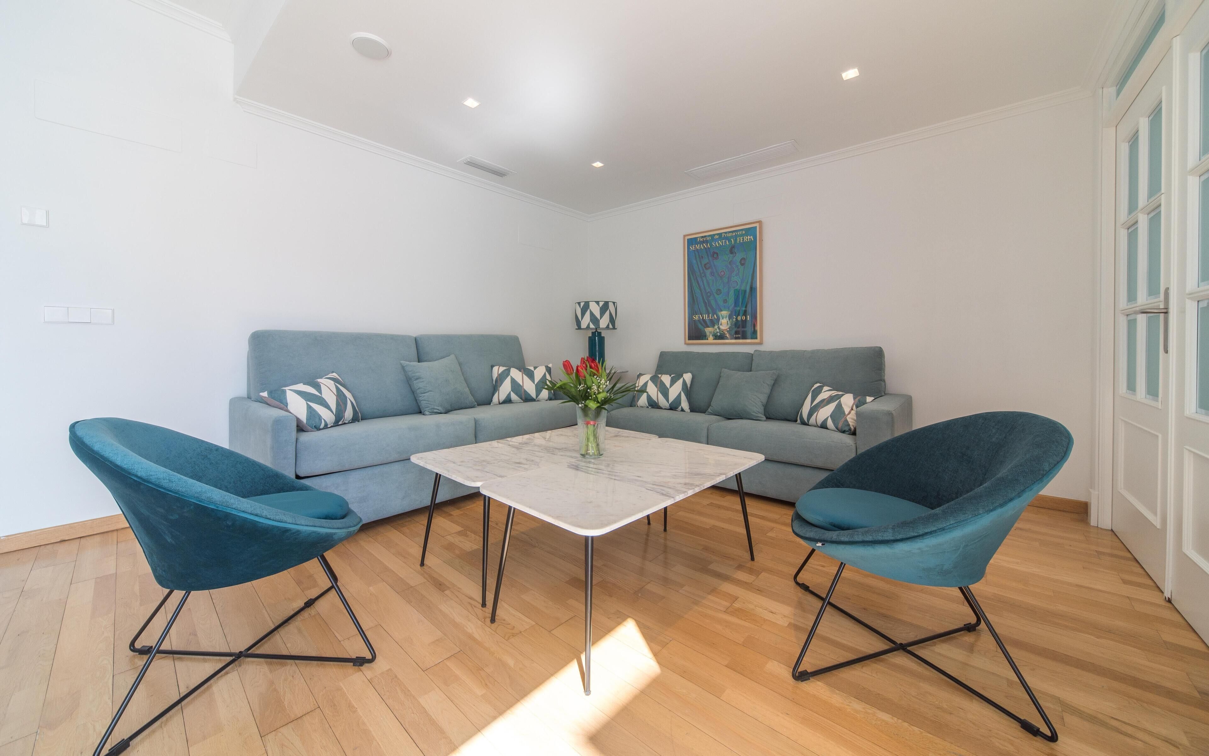 A modern living room with a grey sofa, two blue armchairs, a white coffee table, and a decorative vase with flowers.
