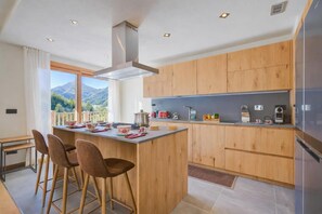 Une cuisine moderne avec des meubles en bois, un îlot central doté de tabourets, une grande fenêtre offrant une vue sur les montagnes et une cuisinière encastrée.