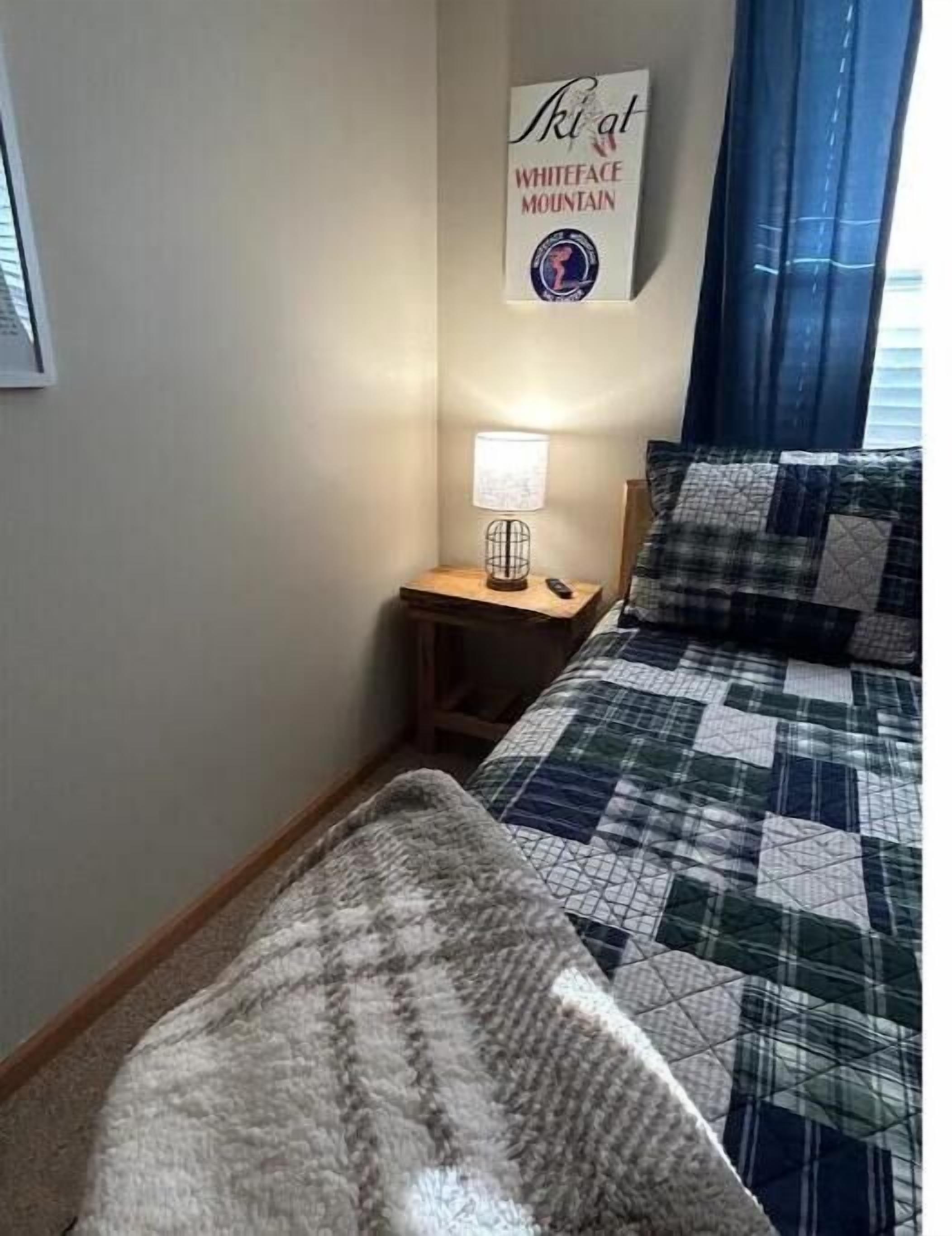 A bedroom with a bed covered in a green and white quilt, a bedside lamp, and a sign on the wall that reads "Ski at Whiteface Mountain."