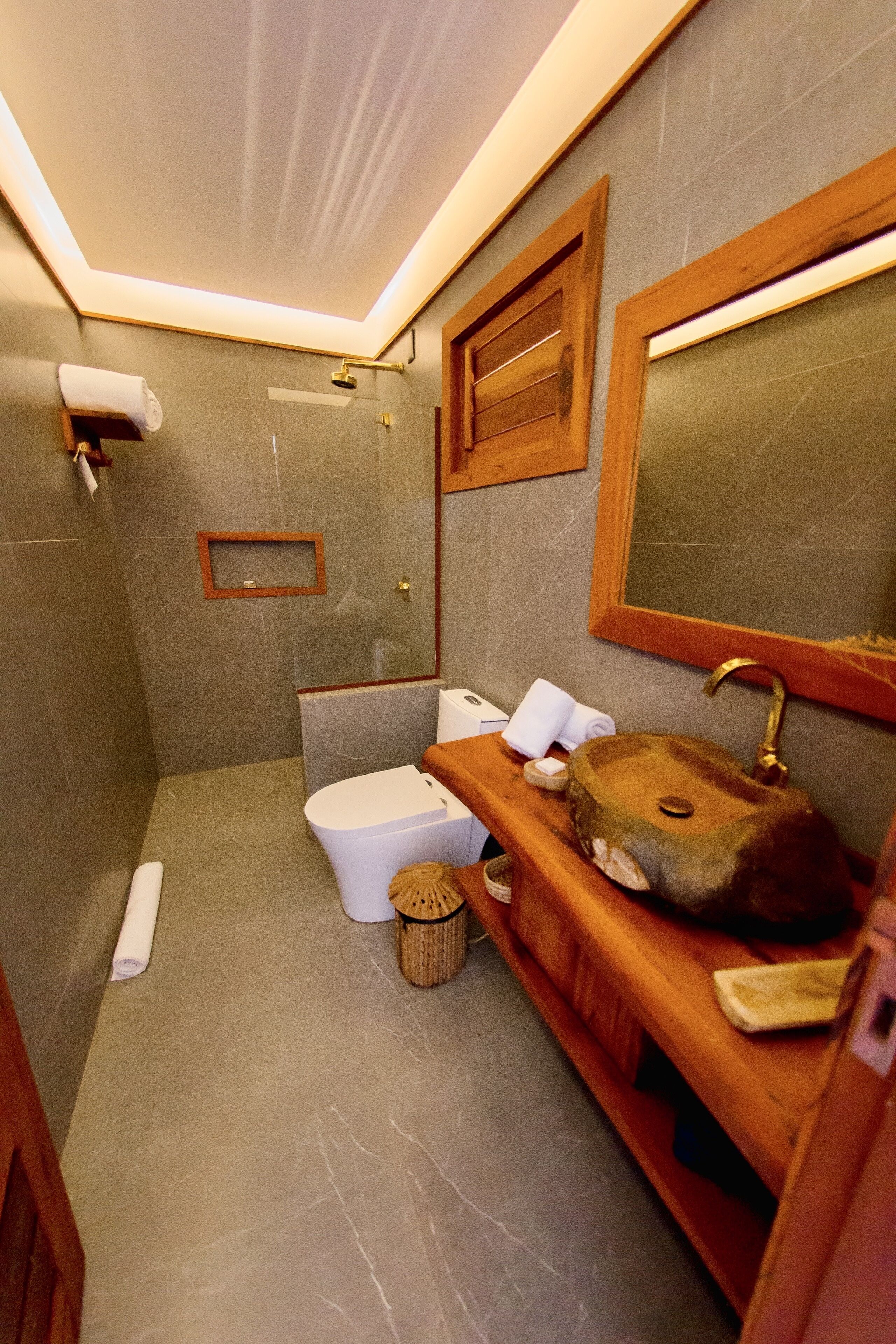 A compact bathroom with a marble floor, a wooden vanity, a white toilet, a glass shower enclosure, and a mirror above the sink.