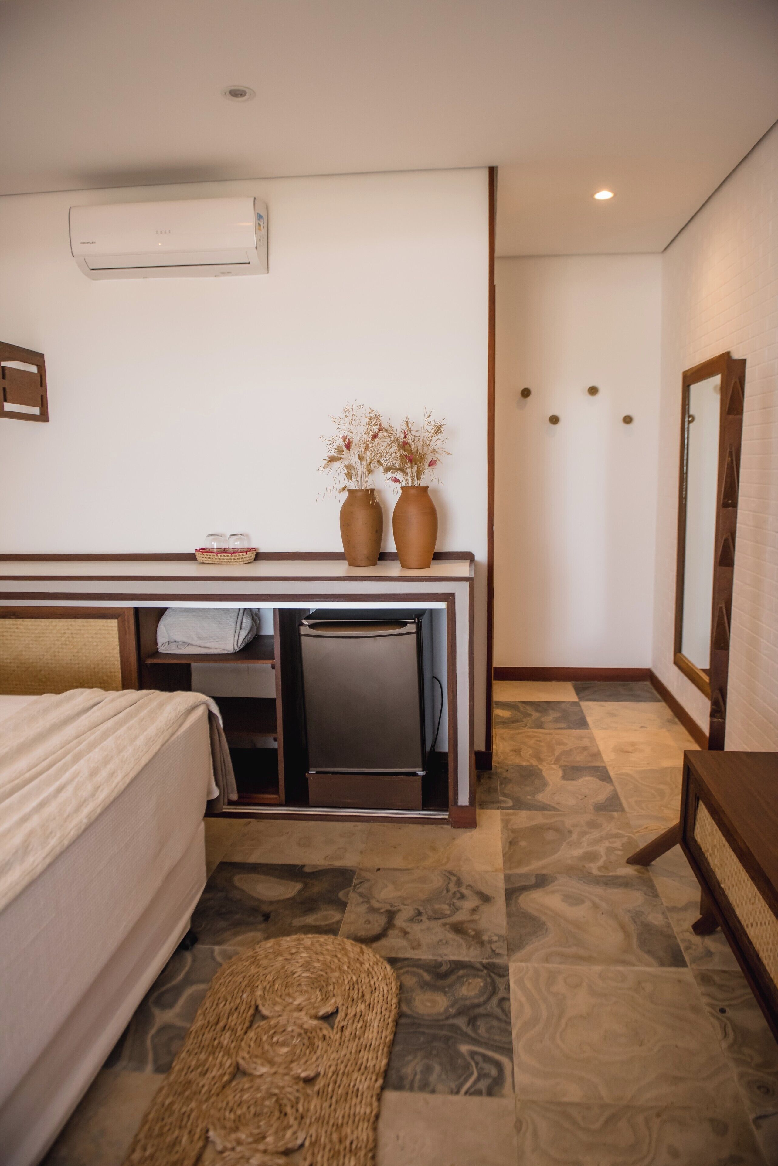 Ein Hotelzimmer mit Bett, Schreibtisch mit Computer, kleinem Tisch, einer Vase mit getrockneten Blumen und einem Spiegel.