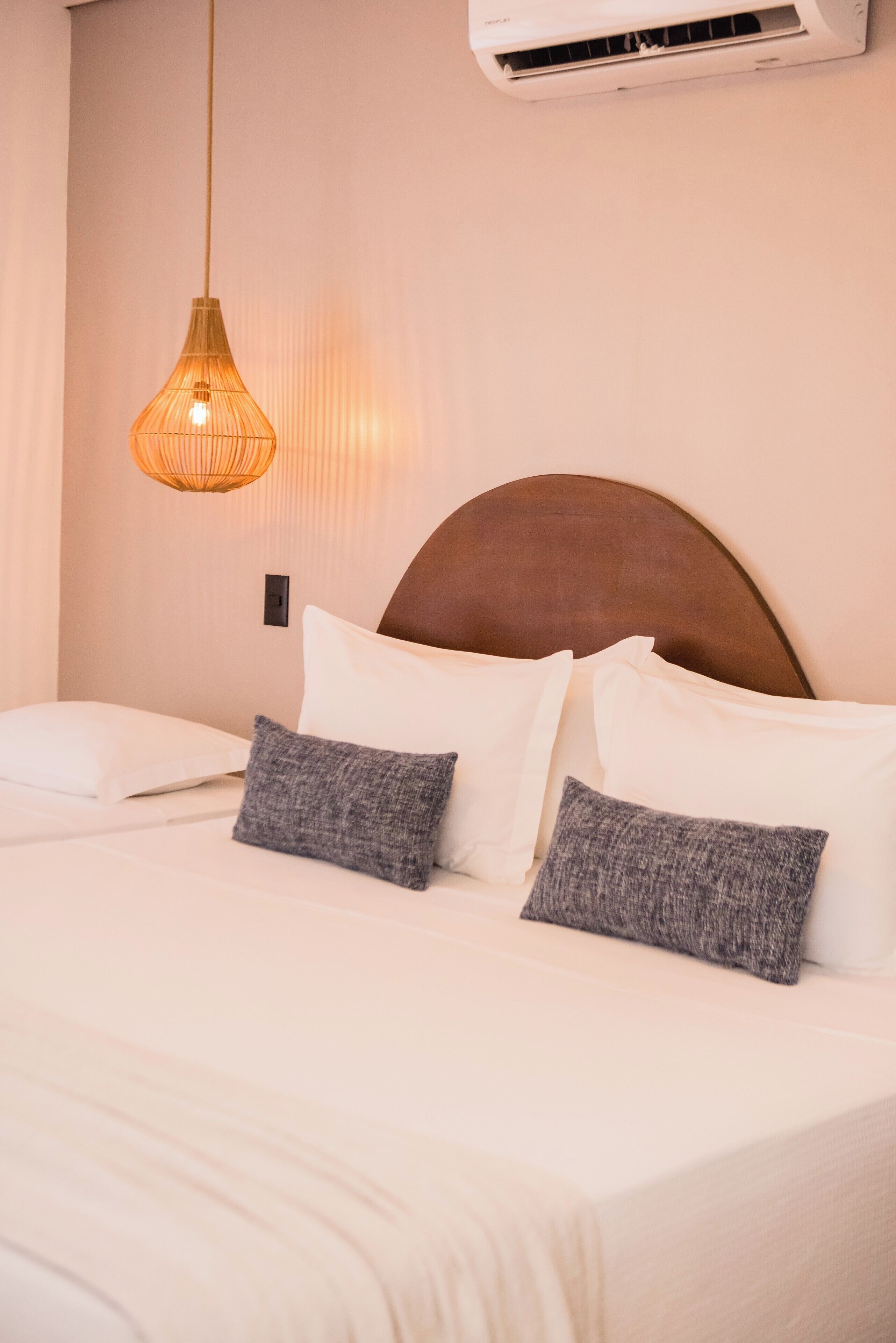 A neatly made bed with white and gray pillows, a hanging pendant light, and an air conditioning unit on the wall.