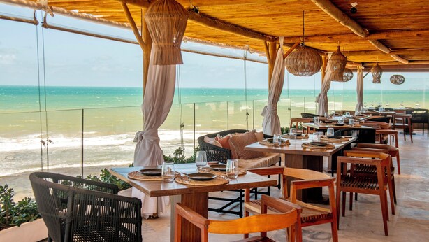 A beachside restaurant with wooden tables and chairs, hanging wicker lamps, and a view of the ocean. - Carlore Hotel Taíba (São Gonçalo do Amarante)