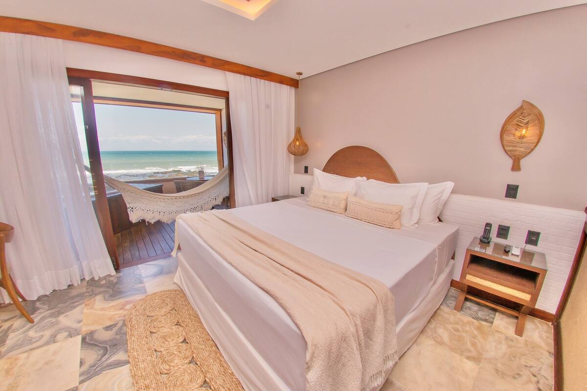 A bedroom with a large bed, a view of the beach, a hammock, and a bedside table with a lamp.
