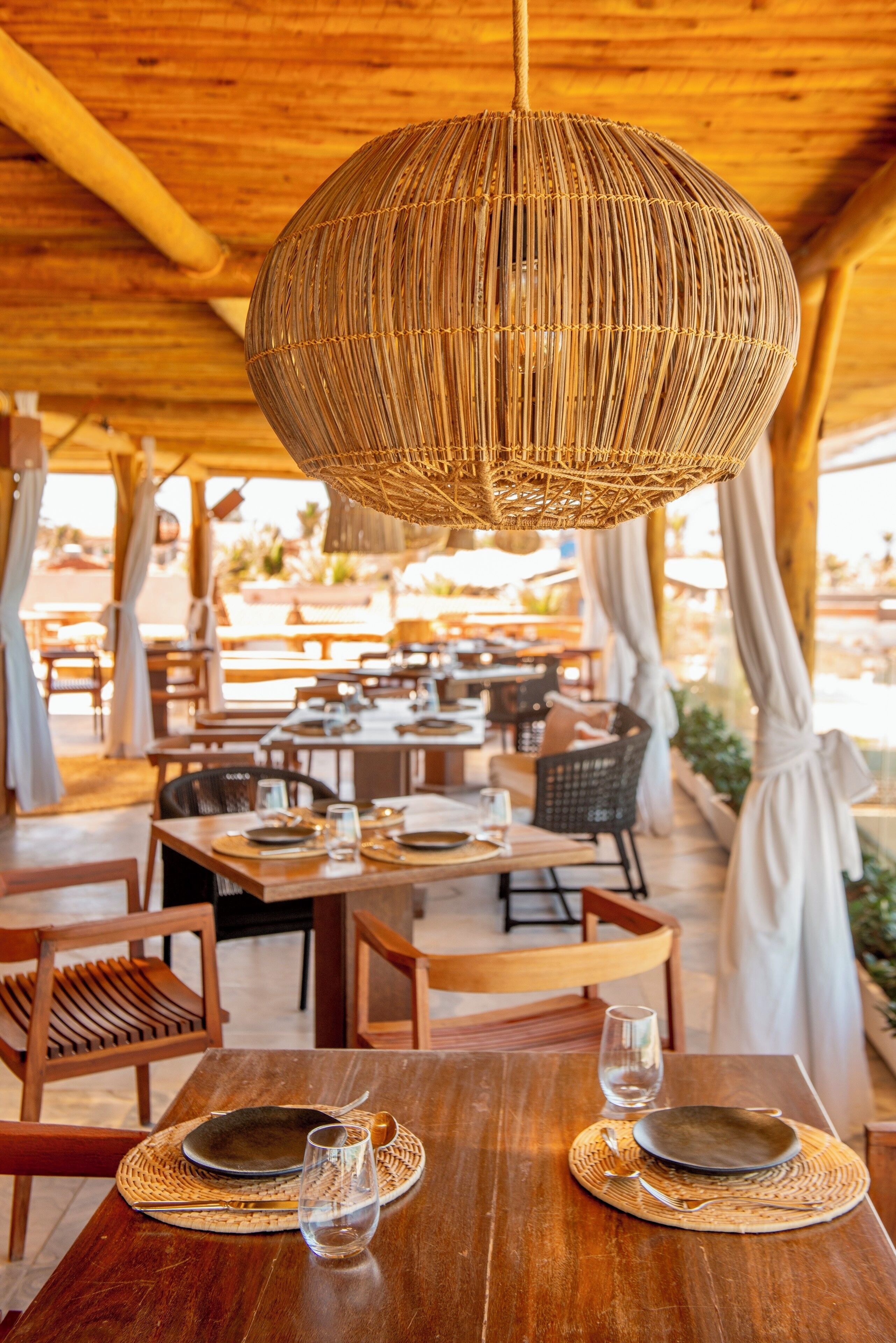 An outdoor dining area with wooden tables and chairs, a woven pendant light, and a view of the surrounding landscape.