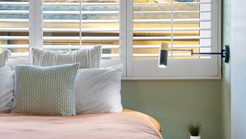 Un lit bien fait, recouvert d’une couvre-lit et de coussins aux couleurs claires, une table de chevet avec une petite plante en pot, et une fenêtre avec des stores blancs et une lampe suspendue.