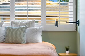 A neatly made bed with a light-colored bedspread and pillows, a bedside table with a small potted plant, and a window with white blinds and a hanging lamp. - Heated salt water pool Spacious 4BR,3BR (Dunedin)