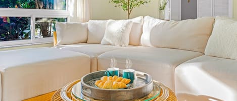 A modern living room with a sectional sofa, a wicker coffee table with croissants and coffee cups, and a view of greenery outside.