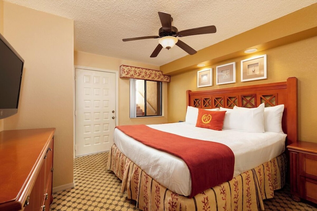 Habitación de hotel con cama, ventilador de techo, cabecera de madera, televisor, puerta y ventana con cortina.