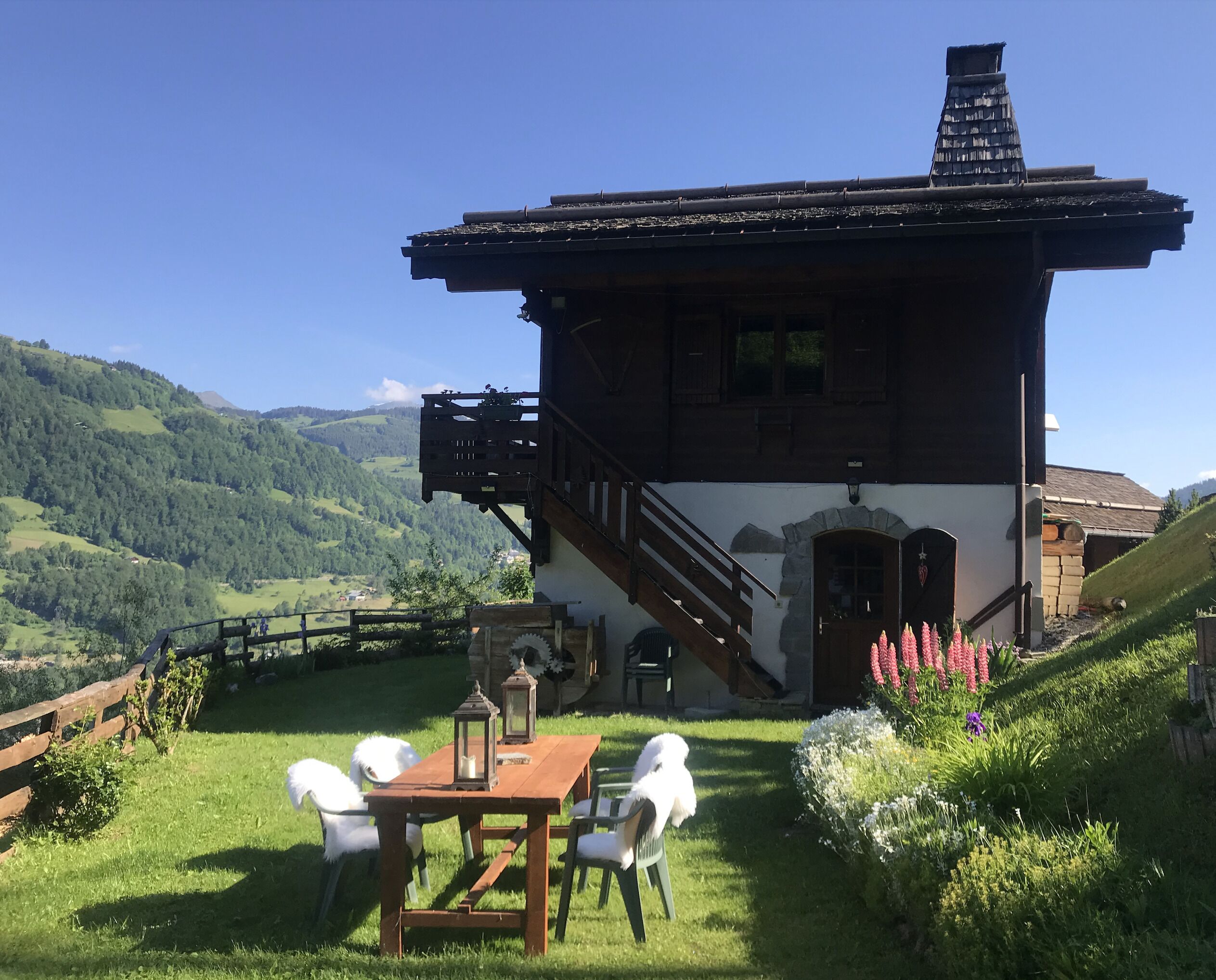 A chalet with a wooden staircase leading to a balcony, surrounded by lush greenery and a mountainous backdrop.