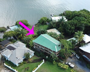 An aerial view of a coastal property with a large house featuring a green roof, surrounded by lush greenery and a dock extending into the water.