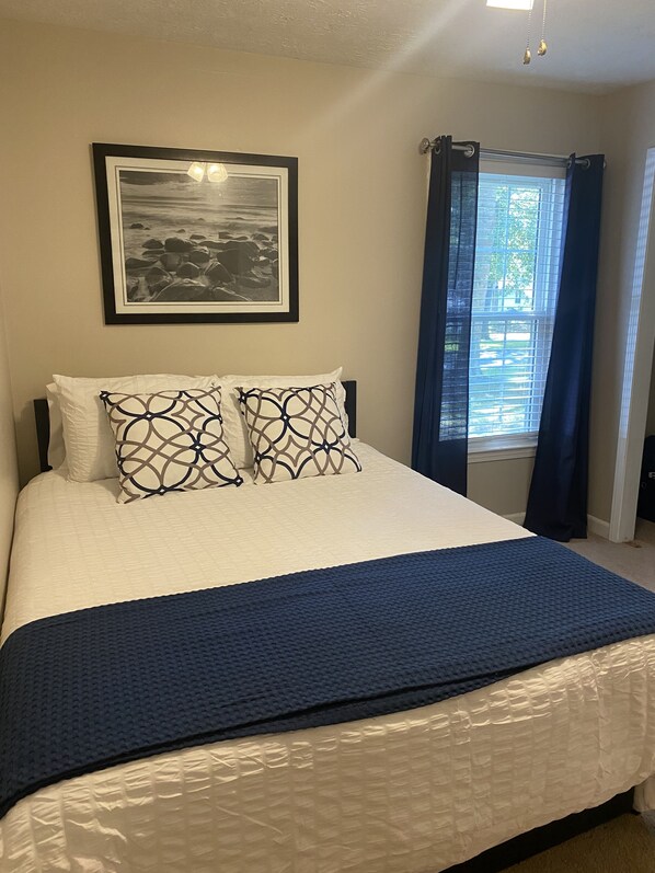 Una cama bien hecha con una colcha blanca y azul, dos almohadas estampadas y un cuadro en la pared.